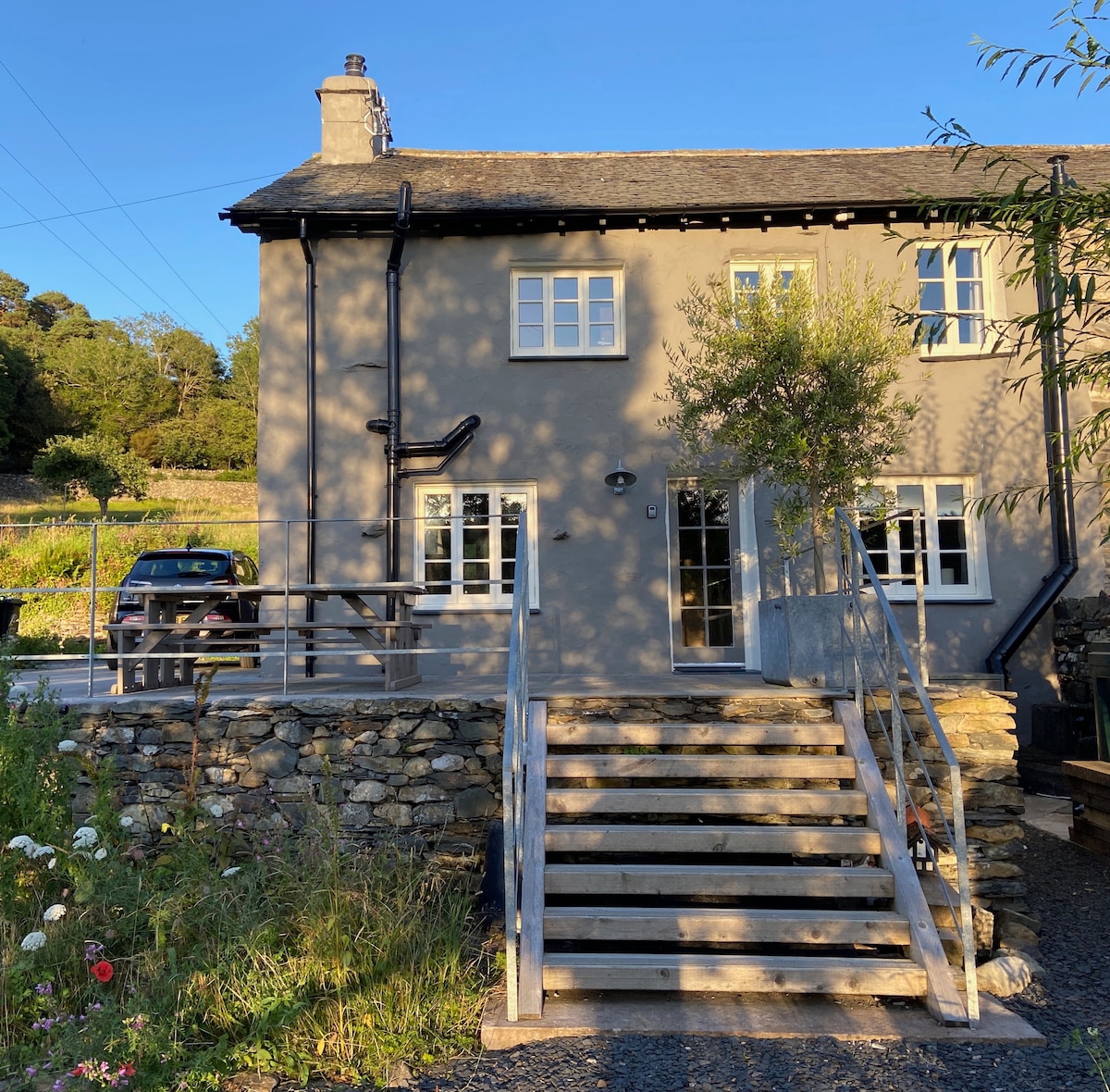 The exterior of a charming two-story cottage is displayed, featuring a prominent set of wooden stairs leading to the entrance. A car is parked nearby, and the surrounding landscape includes greenery and wildflowers, reflecting a peaceful rural setting in the Lake District.