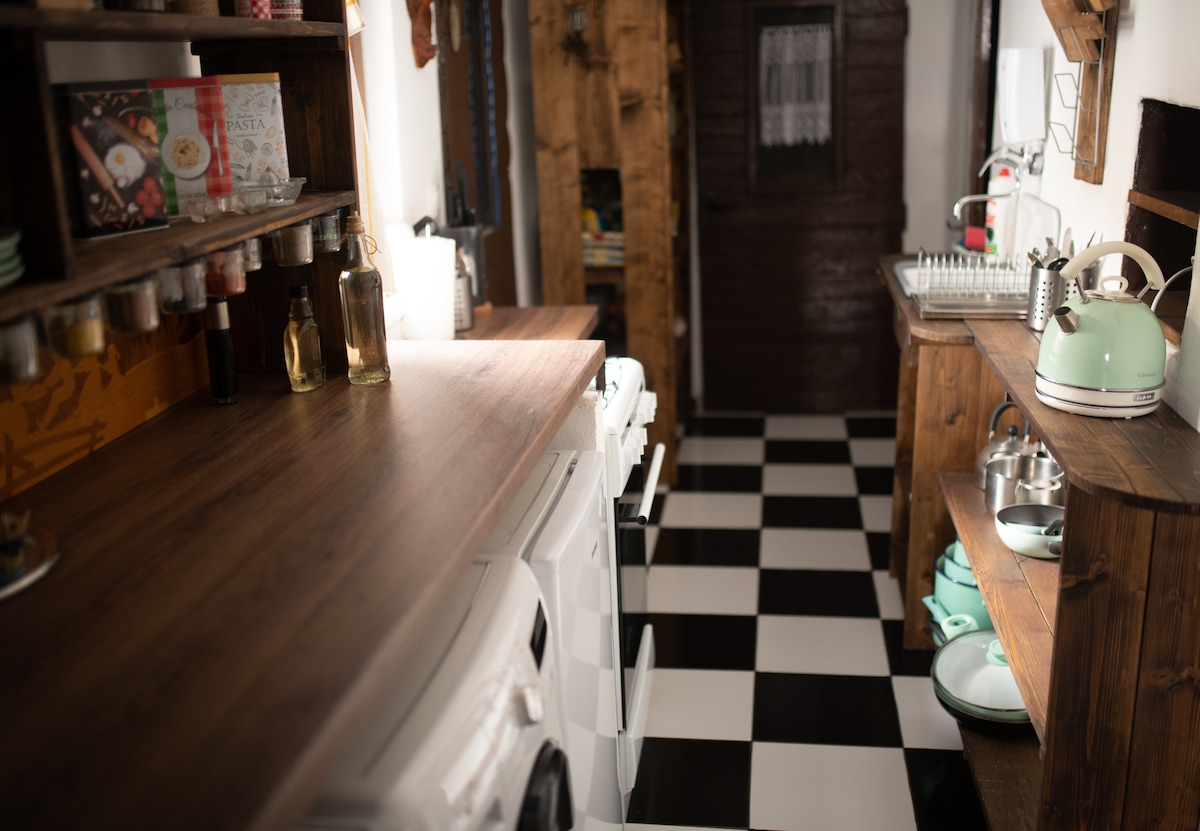 A functional kitchen area features a combination of dark wooden cabinetry and modern appliances. A checkered floor contrasts with the sleek, dark countertop. Cooking essentials are neatly organized on the shelves, with various glass jars and utensils visible, creating an inviting and practical cooking space.