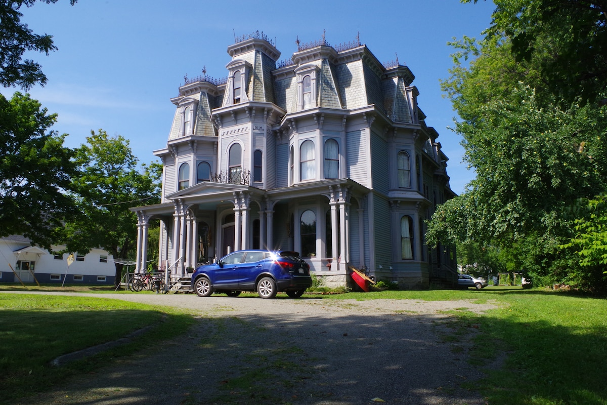 The historic Todd Mansion showcases its elegant architecture, marked by intricate details and a decorative roofline. The tranquil grounds are surrounded by lush greenery, with a gravel driveway accommodating a parked car. The inviting façade features large front pillars and multiple windows.