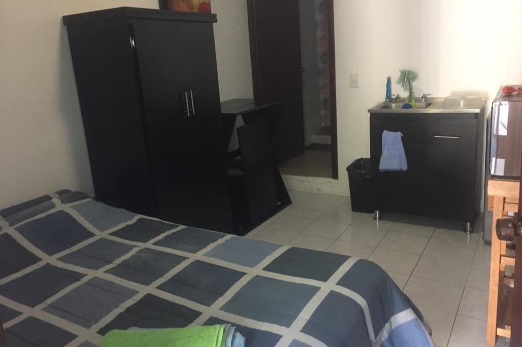 The room contains a spacious bed covered with a patterned blanket. A large wardrobe and a work desk are positioned nearby. A small kitchenette is visible, showcasing a sink, refrigerator, and microwave. Tiled flooring and neutral walls contribute to a clean, functional environment.