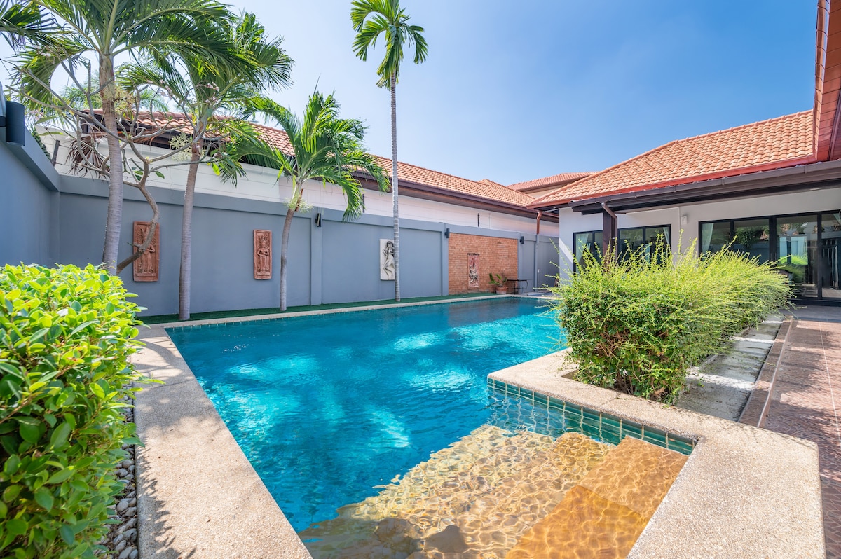 An expansive outdoor pool is framed by lush palm trees and vibrant greenery. The clear blue water reflects sunlight, inviting relaxation. Adjacent to the pool, a spacious terrace offers a comfortable space for lounging and enjoying the outdoors, creating a serene environment.