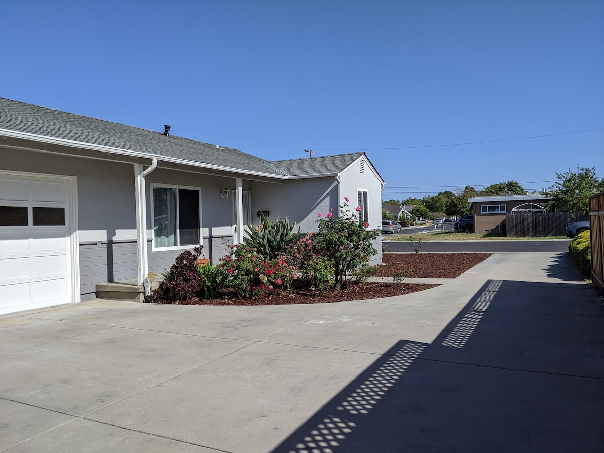 A well-maintained exterior view showcases the front yard of the duplex, featuring gravel beds and blooming shrubs. A driveway leads to a one-car garage on the left, while ample outdoor space is presented for relaxation or play.