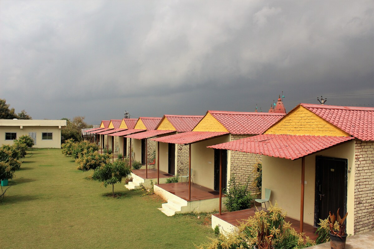 A row of quaint cottages with yellow and red roofs is set against a backdrop of dark clouds. Each cottage features a wooden door and is surrounded by well-maintained greenery, creating a serene and peaceful environment.