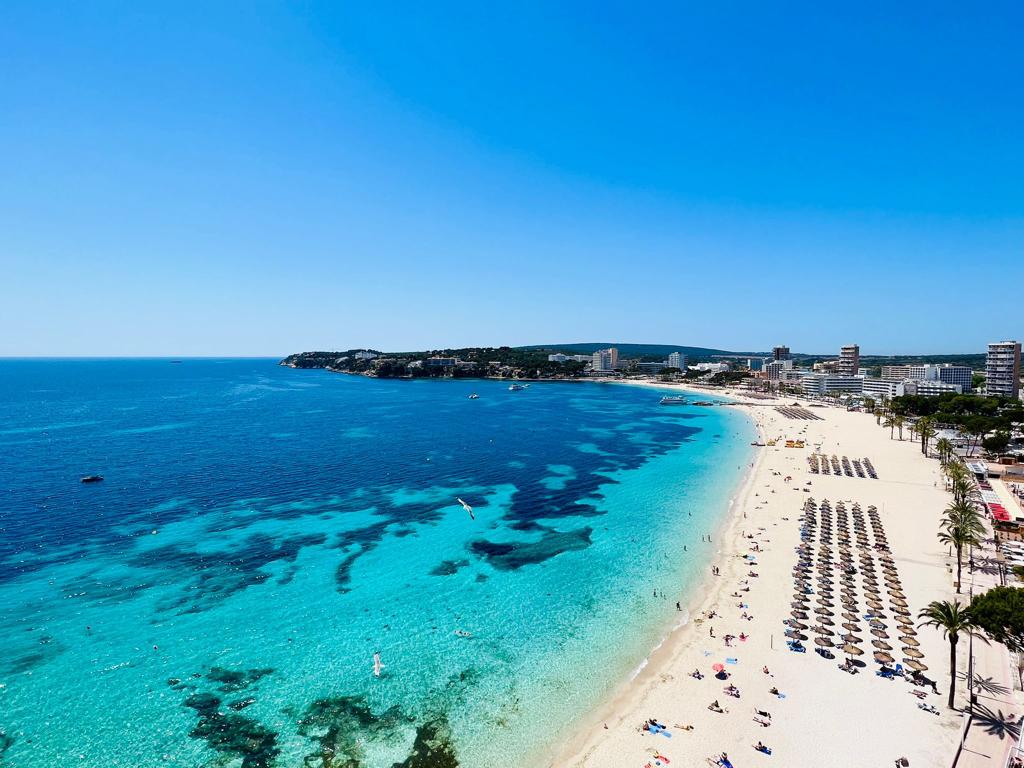 A sweeping view reveals a sandy beach lined with numerous sun loungers under clear blue skies. The turquoise waters gently transition to deeper blues, offering the visual of a calm and inviting shoreline. In the distance, various buildings and palm trees are visible, contributing to the coastal landscape.