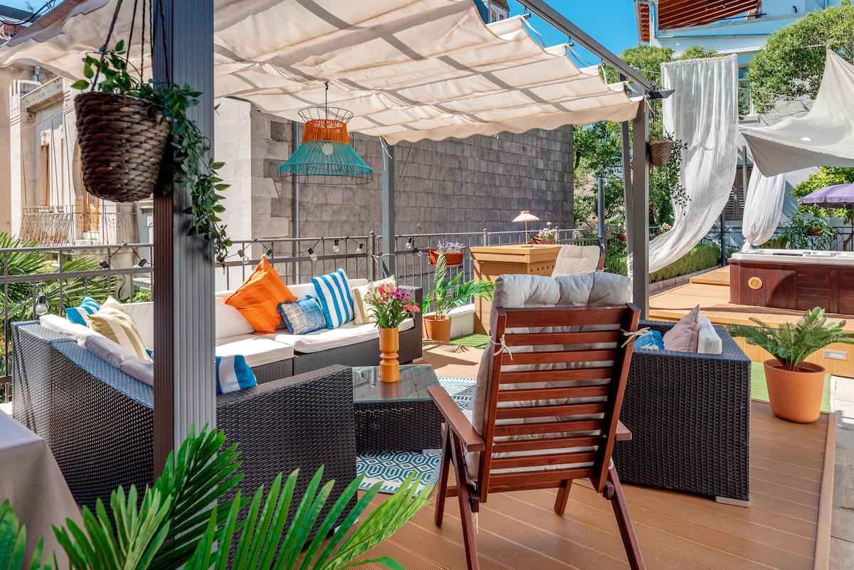 A stylish outdoor lounge area features a combination of comfortable seating with colorful cushions and patterned textiles. Plants are positioned around to enhance greenery. A hot tub and a shaded area are visible in the background, providing a relaxing atmosphere.