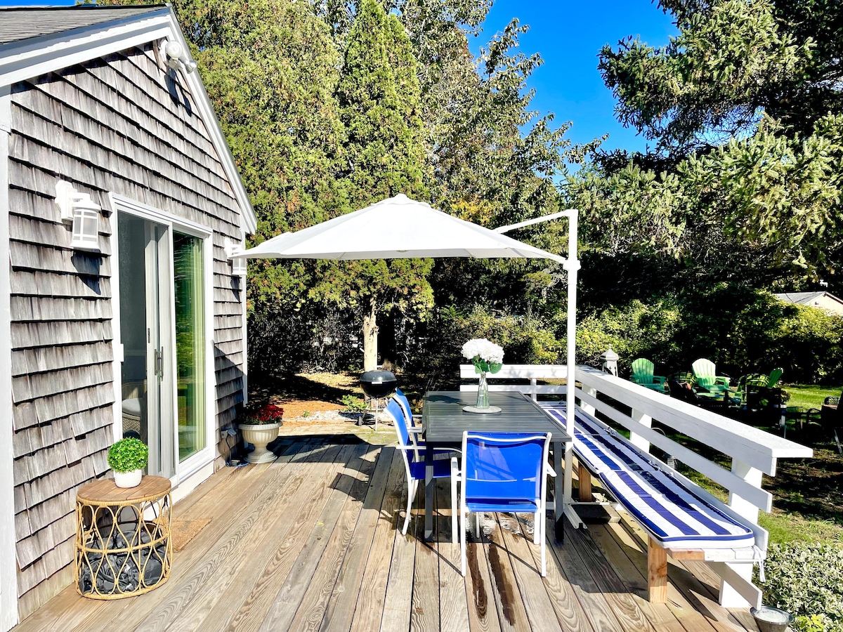An outdoor deck area is shown, featuring a table with chairs, a white umbrella providing shade, and a gas grill set nearby. The wood deck is surrounded by lush greenery, creating a serene environment for dining and relaxing.