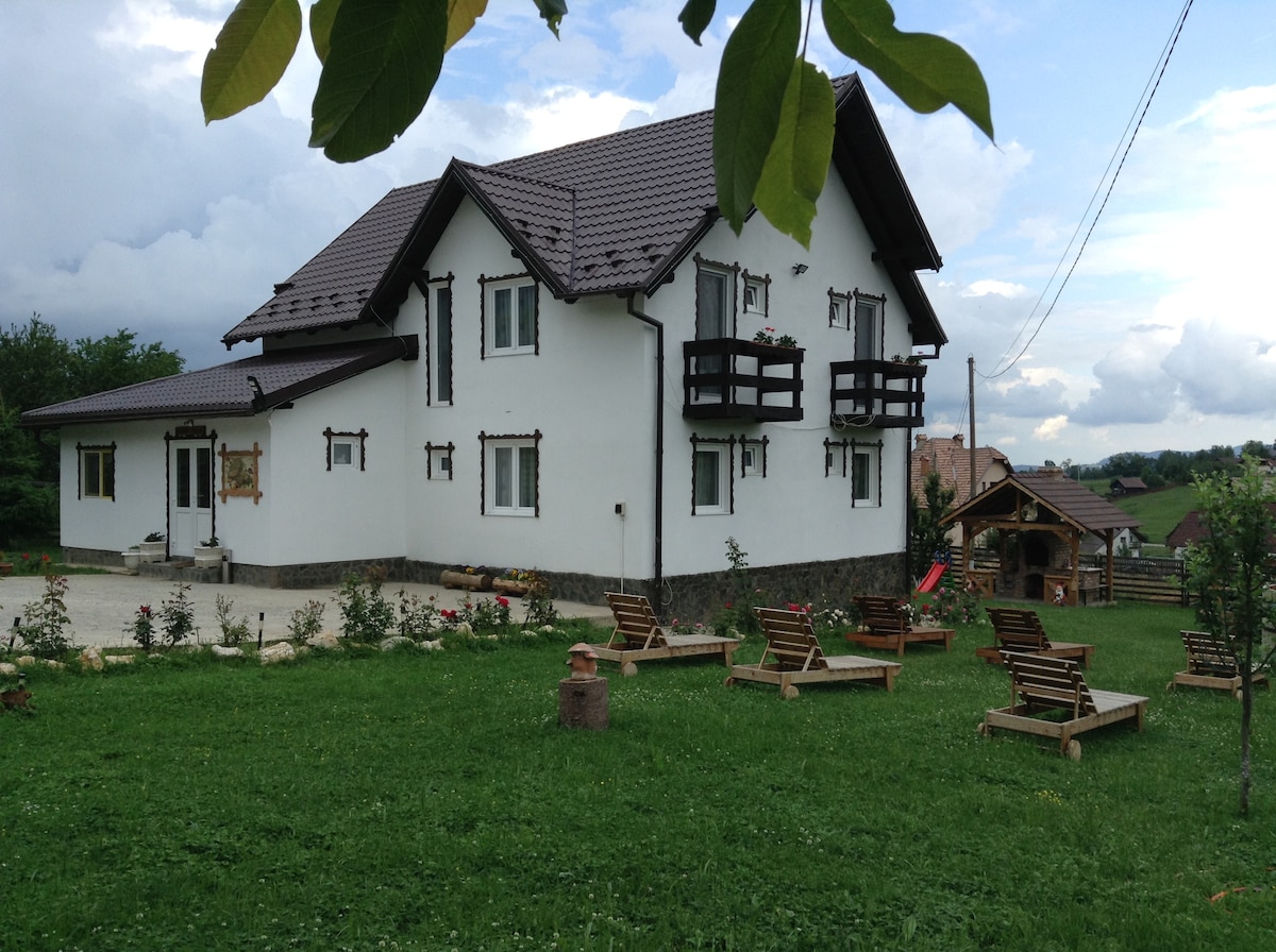 A spacious two-story house with a white exterior is set against a backdrop of greenery. Balconies are featured on the upper levels, and lounge chairs are arranged on the well-maintained lawn. A playground and outdoor seating area are visible, enhancing the serene environment.