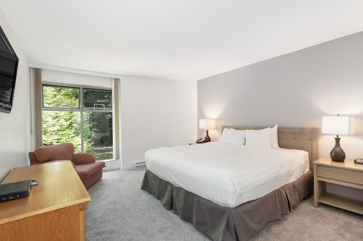 Dormitorio grande tamaño king con TV de pantalla plana, sillón y vista al árbol.