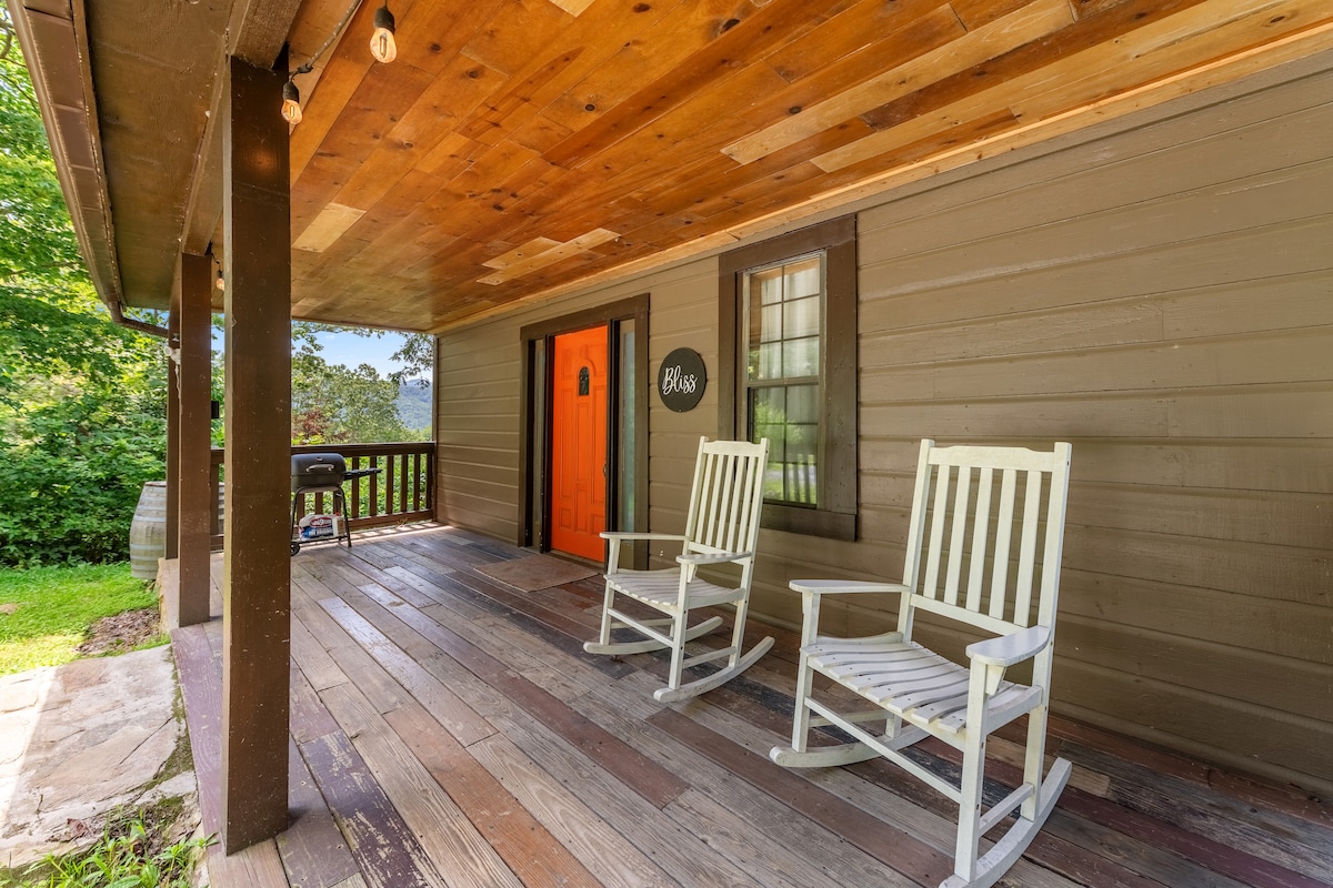 A welcoming porch features two white rocking chairs, inviting relaxation. The entry door is painted a vibrant orange, contrasted against the natural wood tones of the ceiling and floor. Dim lighting from hanging string lights creates a cozy ambiance, surrounded by lush greenery.
