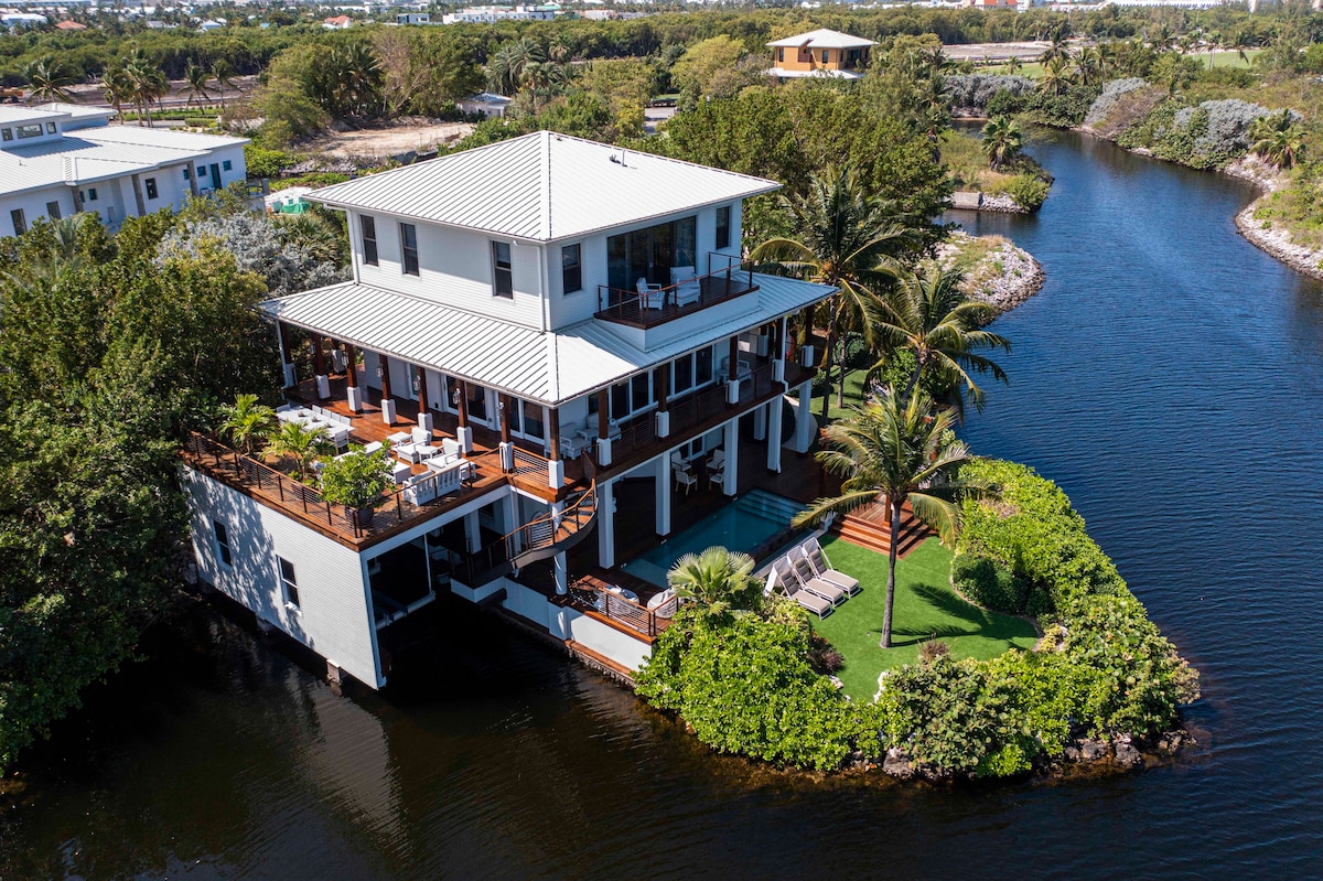 An aerial view of a spacious waterfront home is showcased, surrounded by lush greenery and calm waters. The design features multiple levels with large outdoor decks, sun loungers, and a private pool, all integrated into a serene natural setting.