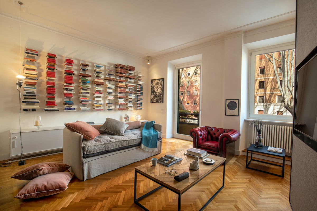 A spacious living area features a large sectional sofa, complemented by decorative pillows and a stylish coffee table. A vibrant bookshelf with numerous titles lines the wall, while a window offers natural light and views of the outside area. A comfortable armchair adds to the inviting atmosphere.