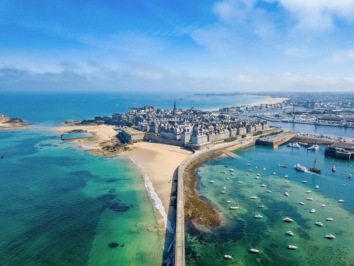 An aerial view captures the historical coastal town with sandy beaches and an array of small boats dotting the turquoise waters. The fortified city, with stone buildings, stretches along the shoreline, showcasing a mix of natural beauty and architectural charm under a clear blue sky.
