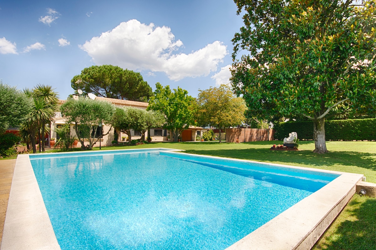 An outdoor swimming pool is set within a spacious garden, surrounded by lush greenery and mature trees. The clear blue water reflects a bright sky and adds an inviting element to the serene setting.