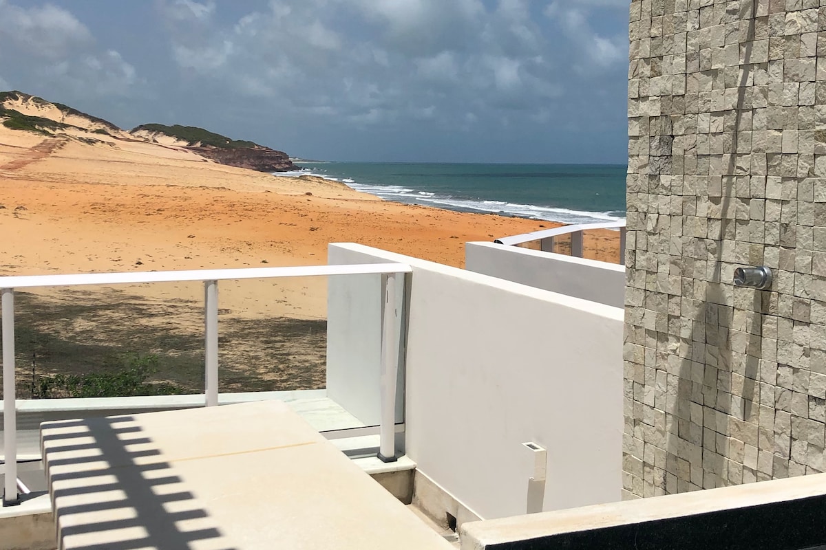 A clear view of the sandy beach and ocean is visible from the terrace. The scene includes the shoreline leading to distant dunes, framed by the modern architectural elements of the terrace. Natural light brightens the area, with clouds scattered across the sky.