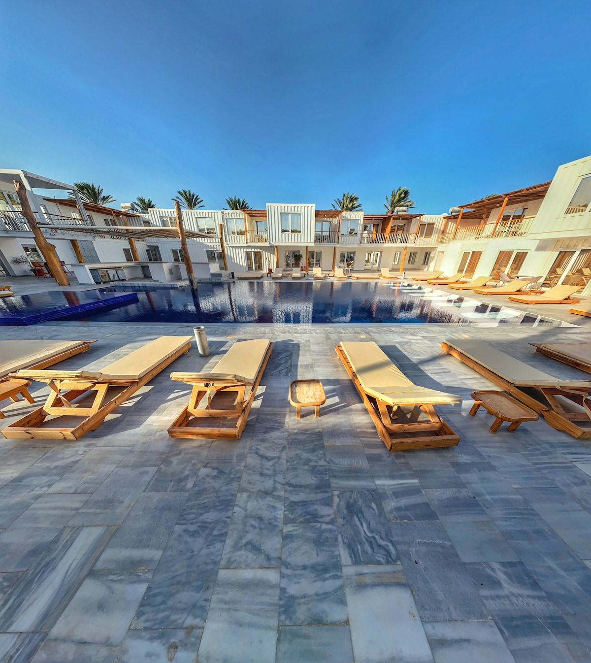 The image displays a vibrant courtyard featuring a large pool surrounded by modern lounge chairs. A series of contemporary buildings are positioned along the perimeter, framed by palm trees under a clear blue sky. Natural stone flooring enhances the overall aesthetic.