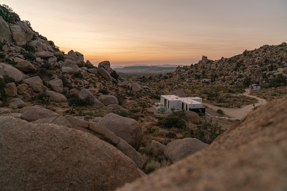 The modern white structure of Sol to Soul is nestled among large granite boulders, surrounded by the rugged desert landscape. A soft sunset casts warm hues across the sky, creating a serene backdrop against the natural terrain.
