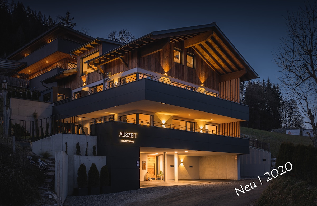 The exterior of a modern apartment building is showcased at dusk, highlighting a combination of wood and sleek design elements. The illuminated entrance features the name 'AUSZEIT', while balconies provide a view of the surrounding natural landscape.