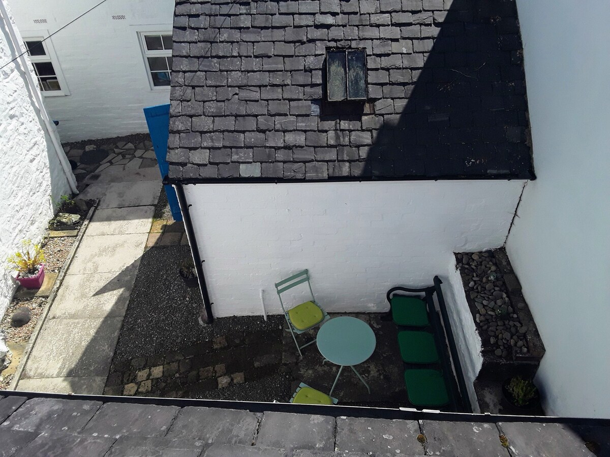 A small outdoor space features a round table with two green chairs against a backdrop of a white wall and a slate-roofed building. The area is paved with stones and complemented by gravel, with a few plants visible around the edges.