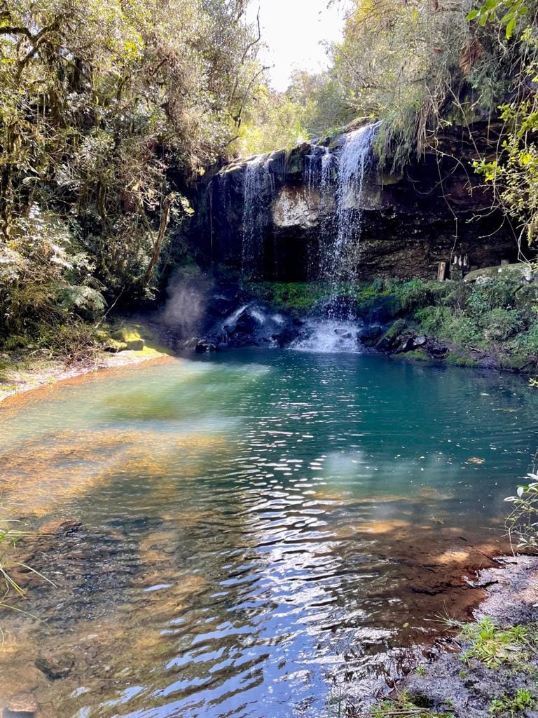 A secluded natural pool is revealed, adorned by a gentle waterfall cascading from rocky ledges. Sunlight filters through surrounding trees, casting dappled reflections on the water's surface. Lush greenery embraces the area, enhancing the tranquil rural setting.