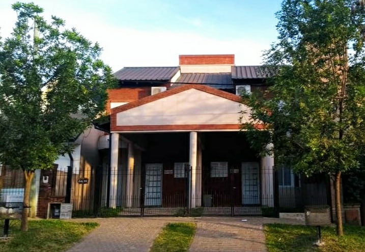 The duplex is situated in a residential tourist area, featuring a welcoming facade with prominent pillars. Lush green trees line the property, providing shade. A well-maintained pathway leads to the entrance, which is enclosed by a decorative black fence.