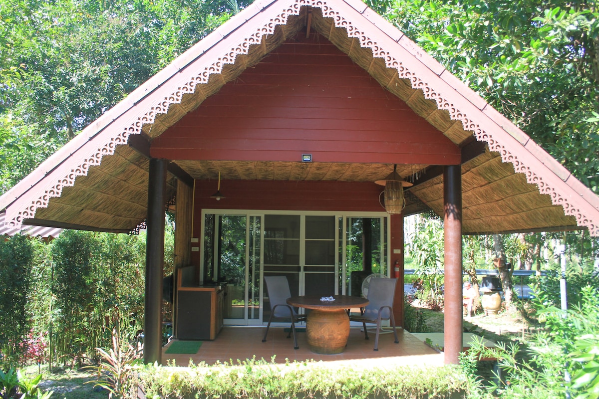 A charming bungalow with a traditional design features a broad porch adorned with two comfortable chairs and a round table. The thatched roof complements the vibrant red exterior, surrounded by lush greenery and tropical plants, providing a serene outdoor setting.