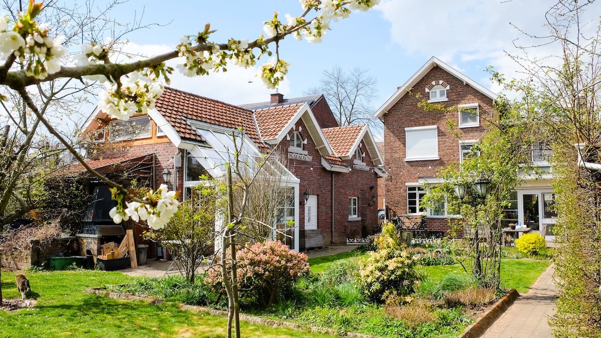 A charming private house is nestled within a vibrant garden, showcasing a blend of brick and traditional architecture. Flowering trees frame the scene, with well-maintained greenery and garden beds surrounding the outdoor space. The structure includes multiple sections, offering a welcoming atmosphere.