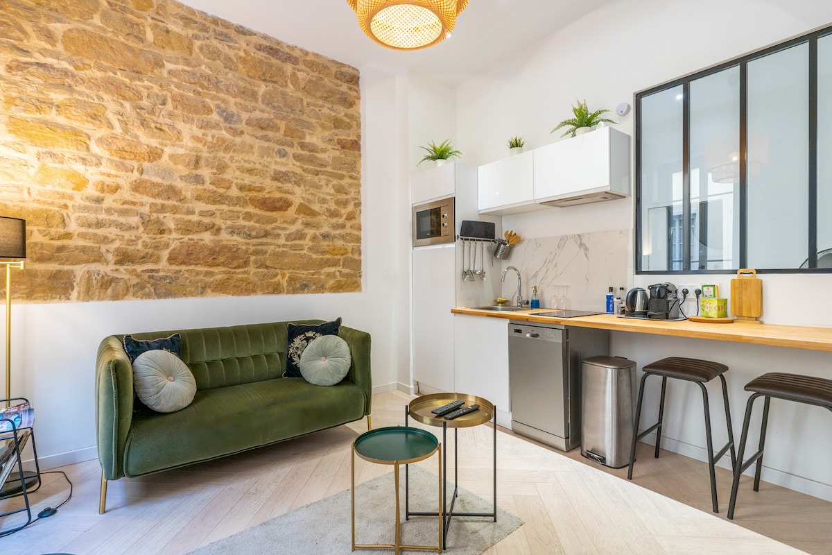 A modern living space features a green velvet sofa with decorative cushions, set against a stylish stone wall. A wooden countertop with bar stools is visible, alongside a kitchenette equipped with appliances. A small side table rests on the layered flooring, completing the inviting atmosphere.