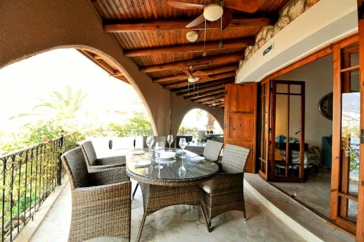 An outdoor dining area features a round table surrounded by six wicker chairs, placed under a wooden ceiling with fans. Arched openings provide views of the surrounding greenery, enhancing the space's airy feel.