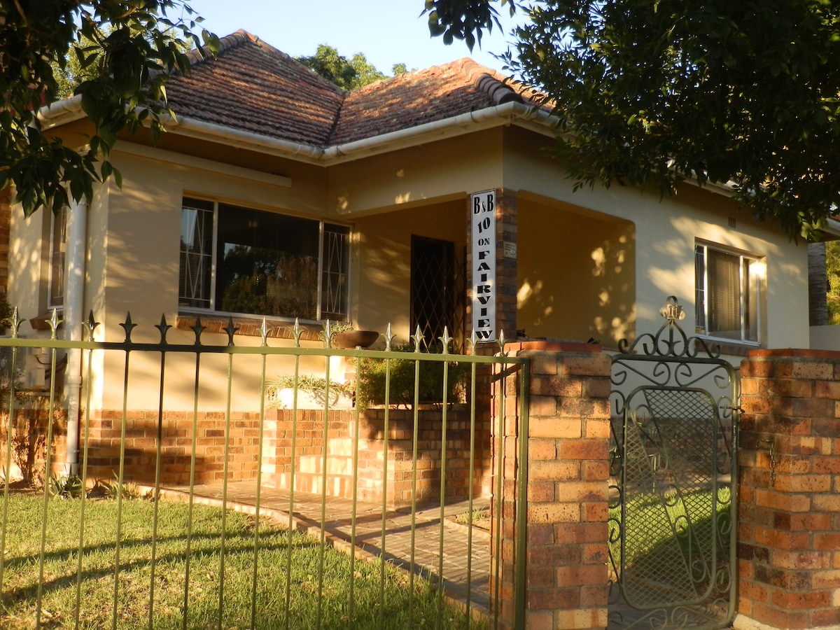 The exterior of a charming home is displayed, featuring a brick and metal gate leading to a path. Large windows allow natural light to illuminate the front rooms. A well-kept lawn surrounds the house, which showcases a sloped roof and a welcoming façade.