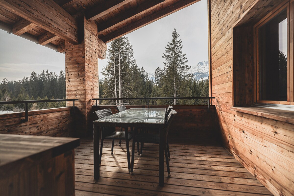 A wooden balcony is furnished with a glass-top dining table and four chairs, surrounded by natural wood paneling. The view showcases tall trees and distant mountains, inviting relaxation and connection with nature.