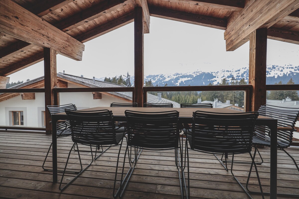 A spacious balcony features a large wooden table surrounded by black metal chairs. The natural wood beams support a sloped roof, and scenic mountain views are visible in the background, creating a serene outdoor ambiance.