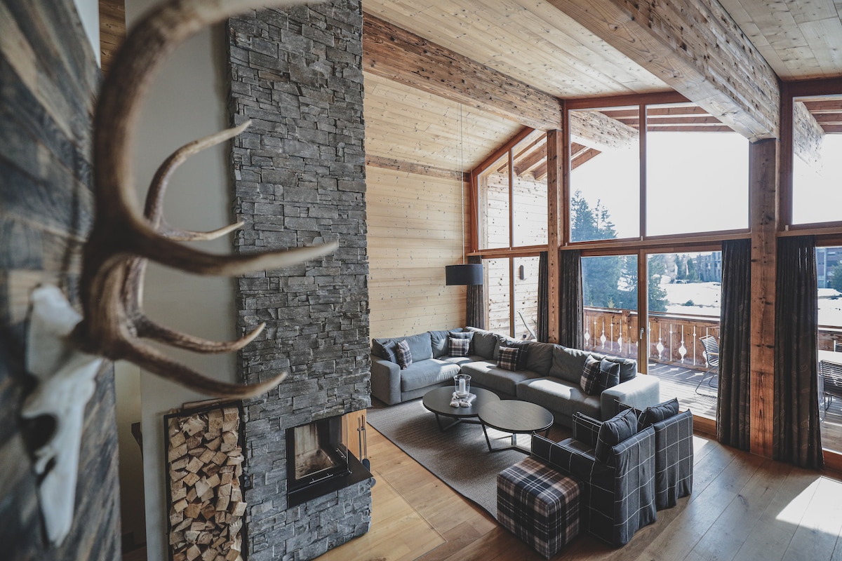 A spacious living area is highlighted by large windows that provide ample natural light and views of the surrounding landscape. A stone fireplace is positioned beside a comfortable seating arrangement, featuring a mix of soft furnishings and wooden accents. The wooden ceiling enhances the chalet's rustic charm.