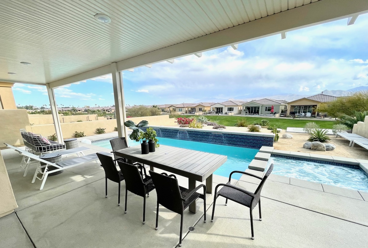 A spacious patio offers a dining table surrounded by black chairs, adjacent to a tranquil salt-water pool. Sun loungers are positioned nearby, while desert landscaping and distant mountain views create a serene backdrop under a partly cloudy sky.
