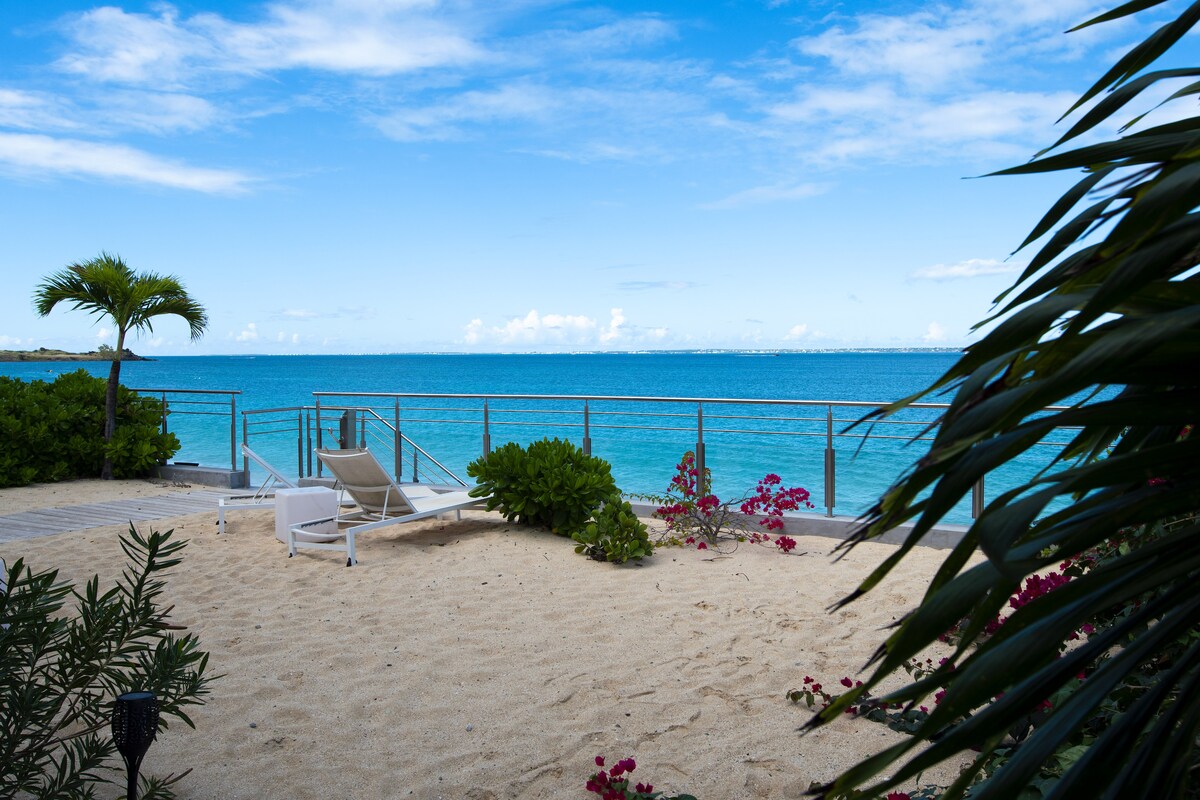 A serene beachfront view showcases gentle waves lapping against the shore, with a partially covered sandy area adorned with lush greenery and vibrant flowers. Two lounge chairs are positioned on the sand, inviting relaxation by the water's edge, framed by palm fronds and coastal plants.