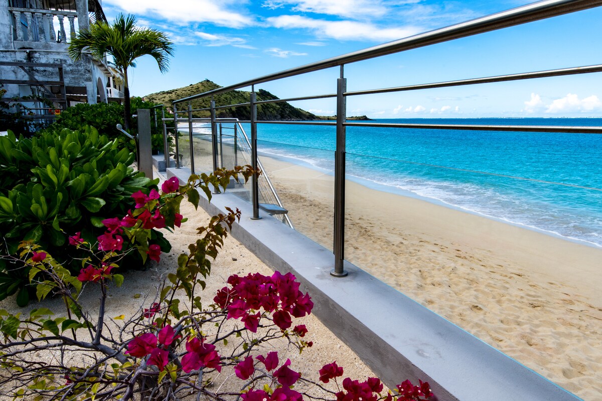 A serene beachfront view is captured, featuring gentle waves lapping against a sandy shore. A low railing lines the deck, complemented by vibrant pink bougainvillea blooms, while lush greenery is seen nearby. The sky showcases scattered clouds above the clear blue water.