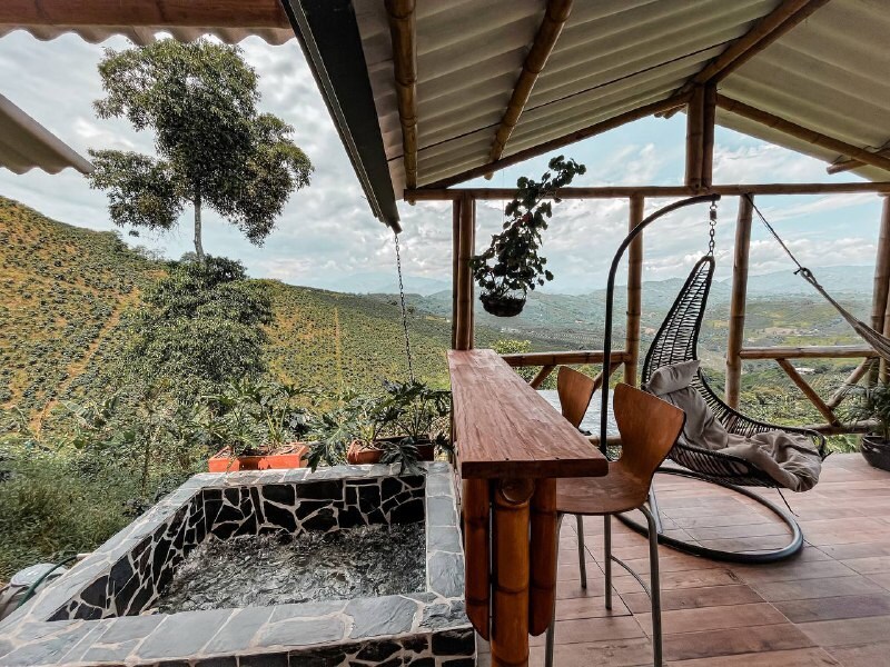 An outdoor balcony features a small jacuzzi alongside a wooden bar table. A stylish hanging chair is positioned nearby, offering views of the lush coffee landscape and distant hills, with potted plants adding a natural touch to the space.