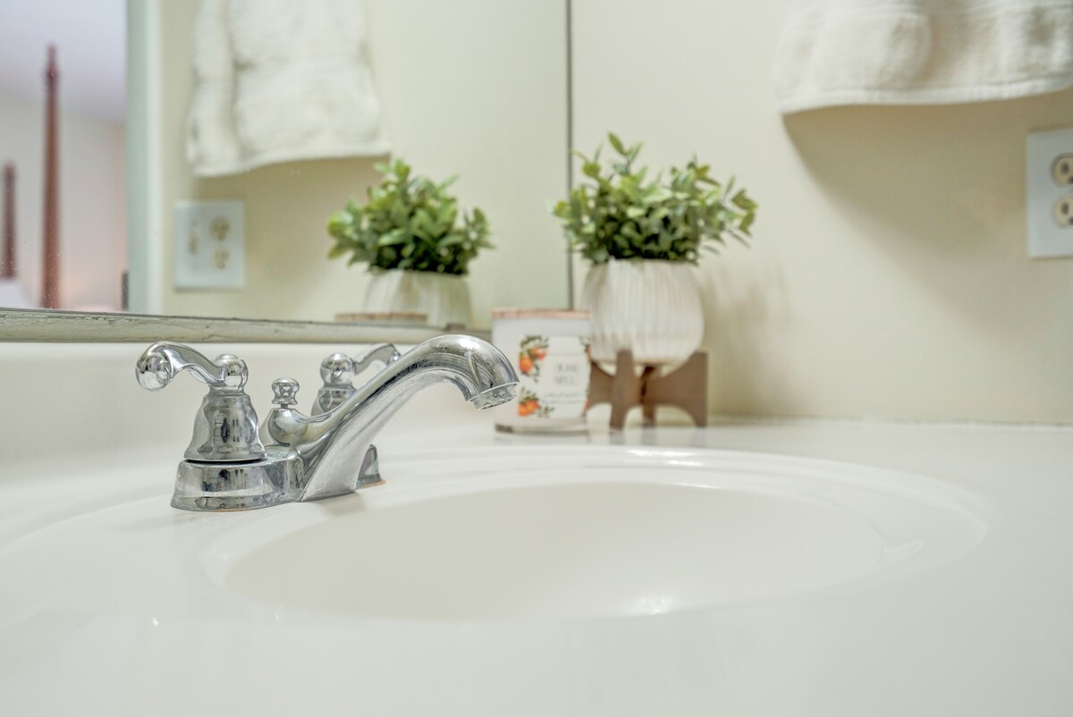 A clean sink area features a polished chrome faucet, positioned on a white countertop. A small potted plant adds a touch of greenery, while toiletries are neatly arranged in the background, contributing to a tidy and comfortable bathroom space.
