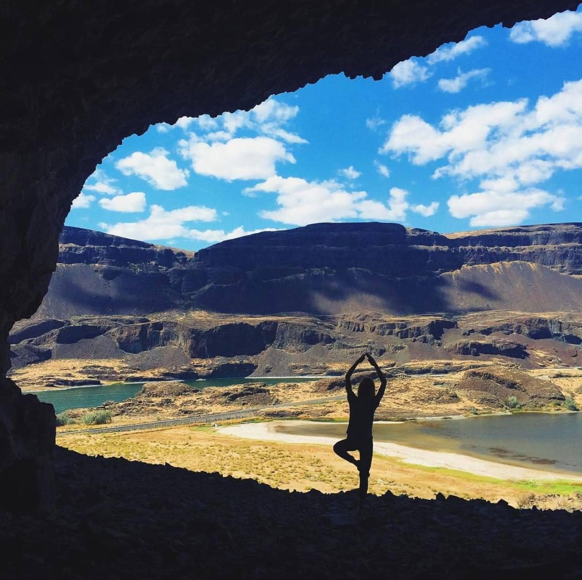 A view from inside a cave showcases a silhouetted figure practicing yoga. The landscape outside reveals a river winding through the valley, framed by rocky cliffs and bright blue skies dotted with clouds.