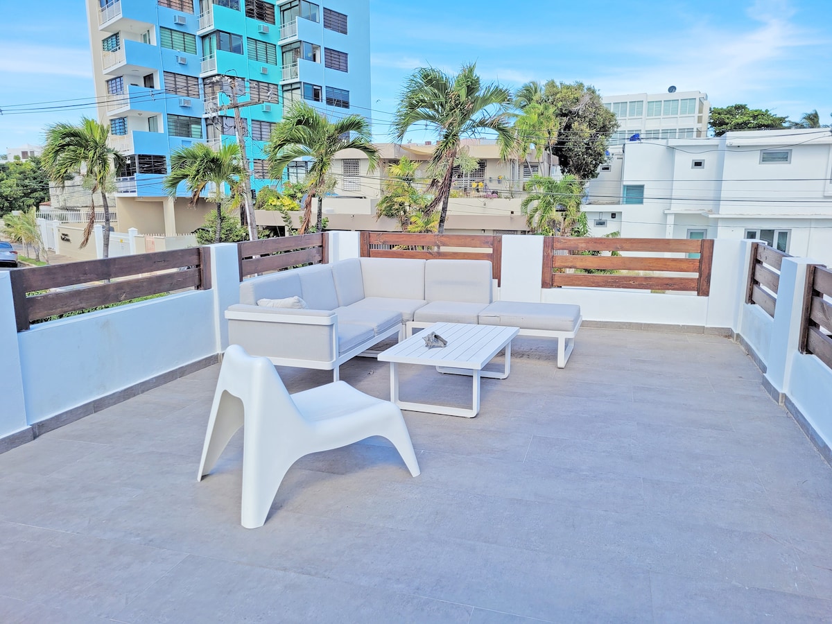 A spacious terrace is adorned with modern outdoor seating, including a sectional sofa and a coffee table. The area is surrounded by a low wooden railing, with a backdrop of palm trees and nearby buildings creating a leisure-friendly environment.
