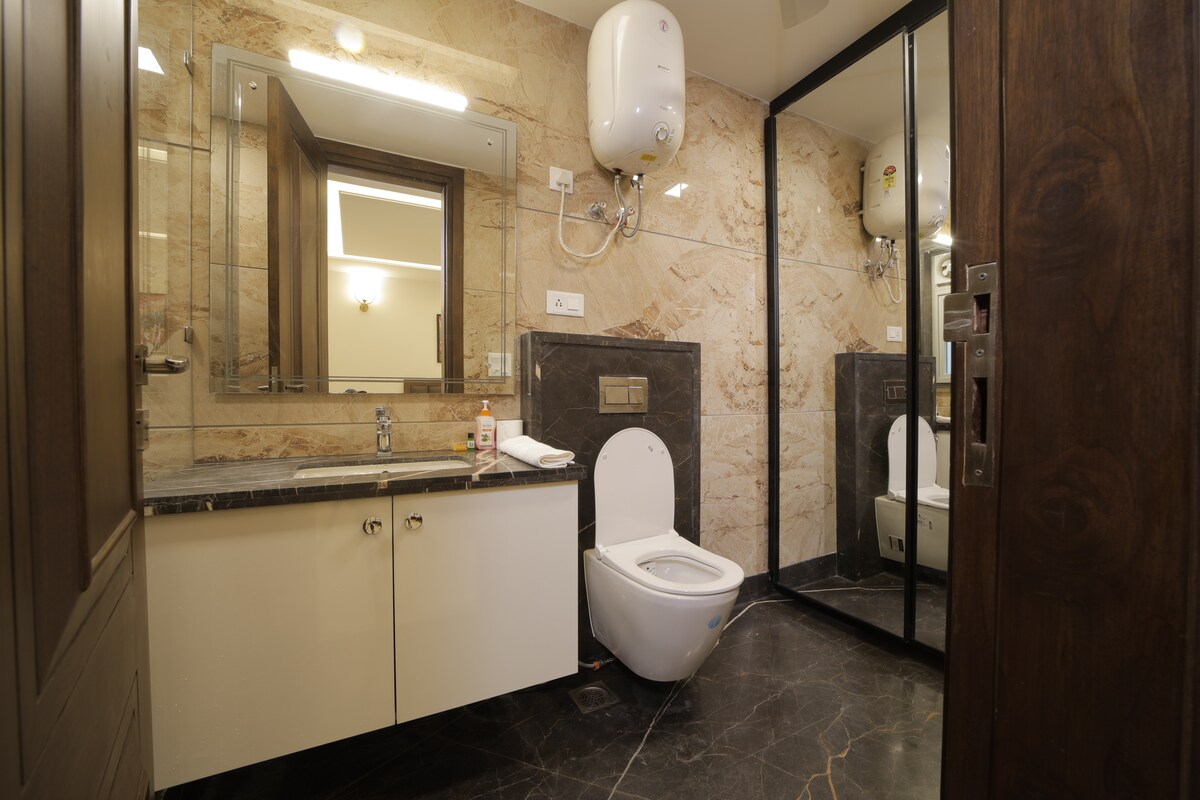 A modern bathroom featuring marble walls and dark stone flooring. A white toilet is positioned next to a sleek vanity with a washbasin. A large mirror and necessary toiletries are visible, along with an electric water heater mounted on the wall.
