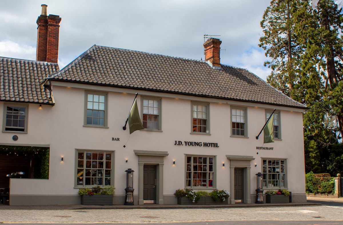 The exterior of J.D. Young Hotel showcases a charming two-story structure with a pitched roof and traditional brick chimneys. Large windows are framed with greenery, and flags are displayed from the front, indicating the bar and restaurant services available.