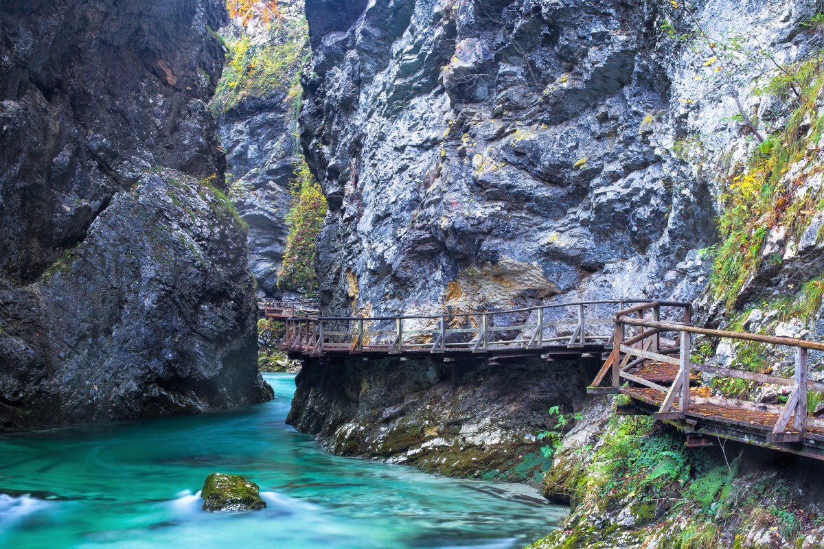 A wooden walkway meanders through a narrow gorge, flanked by towering rocky cliffs. Crystal-clear turquoise water flows gently below, contrasting with the rugged textures of the stone. The lush greenery adds a vibrant touch to the natural scenery.