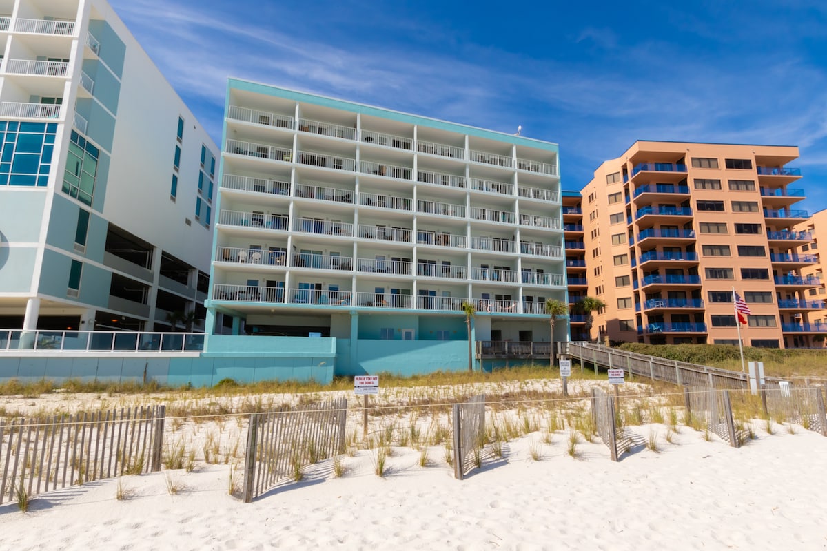 The beachfront property is set against a bright blue sky, showcasing a modern three-story building with spacious balconies. Sandy dunes and beach vegetation lead up to the building, emphasizing its proximity to the shore. Nearby structures exhibit varying colors and architectural styles.