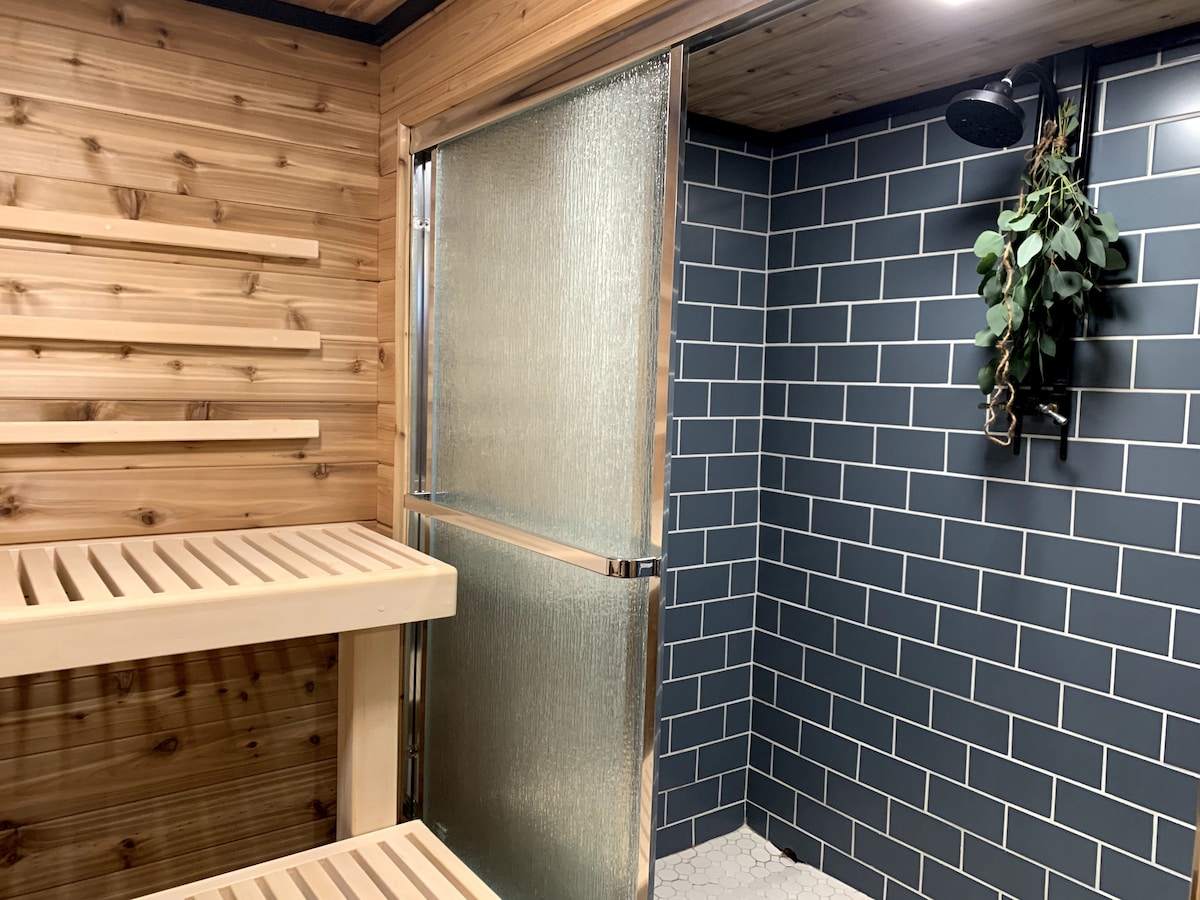 A newly installed cedar sauna offers a serene retreat with wooden slats lining the walls and a sleek tiled shower area nearby. Glass doors provide access to the shower, complemented by a hanging plant for a touch of nature.