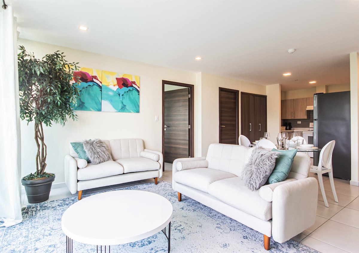 The living area features two light-colored sofas adorned with textured throw pillows. A circular coffee table sits centrally on a soft area rug. Bright artwork hangs on the wall, and the open kitchen is visible through the doorway, ensuring a spacious and inviting atmosphere.