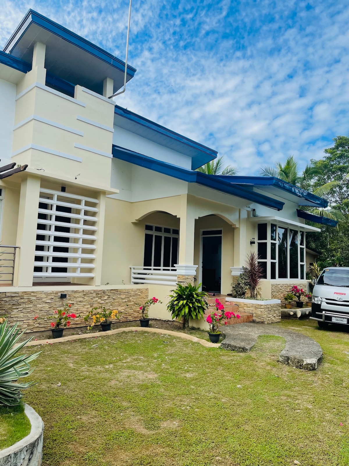 A newly renovated exterior of a house is shown, featuring light-colored walls and a contrasting blue roof. Lush greenery surrounds the entrance, with vibrant flowering plants adding color to the landscape. A paved pathway leads to the front door, enhancing the inviting atmosphere.