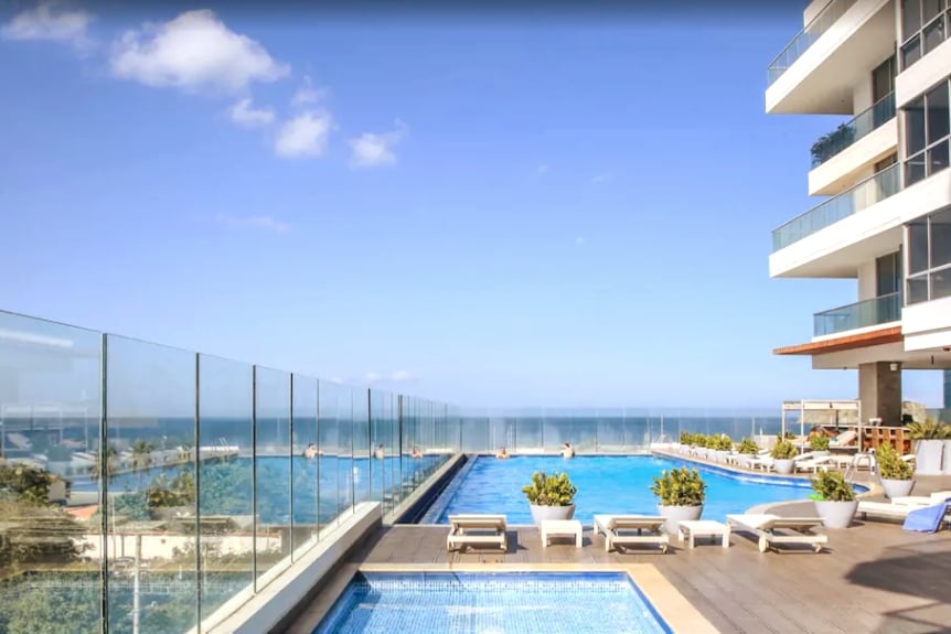 A spacious outdoor pool area is visible, featuring sun loungers arranged around the water. A glass barrier offers unobstructed views of the ocean, enhancing the tranquil atmosphere. The sky above is clear, allowing natural light to illuminate the scene.