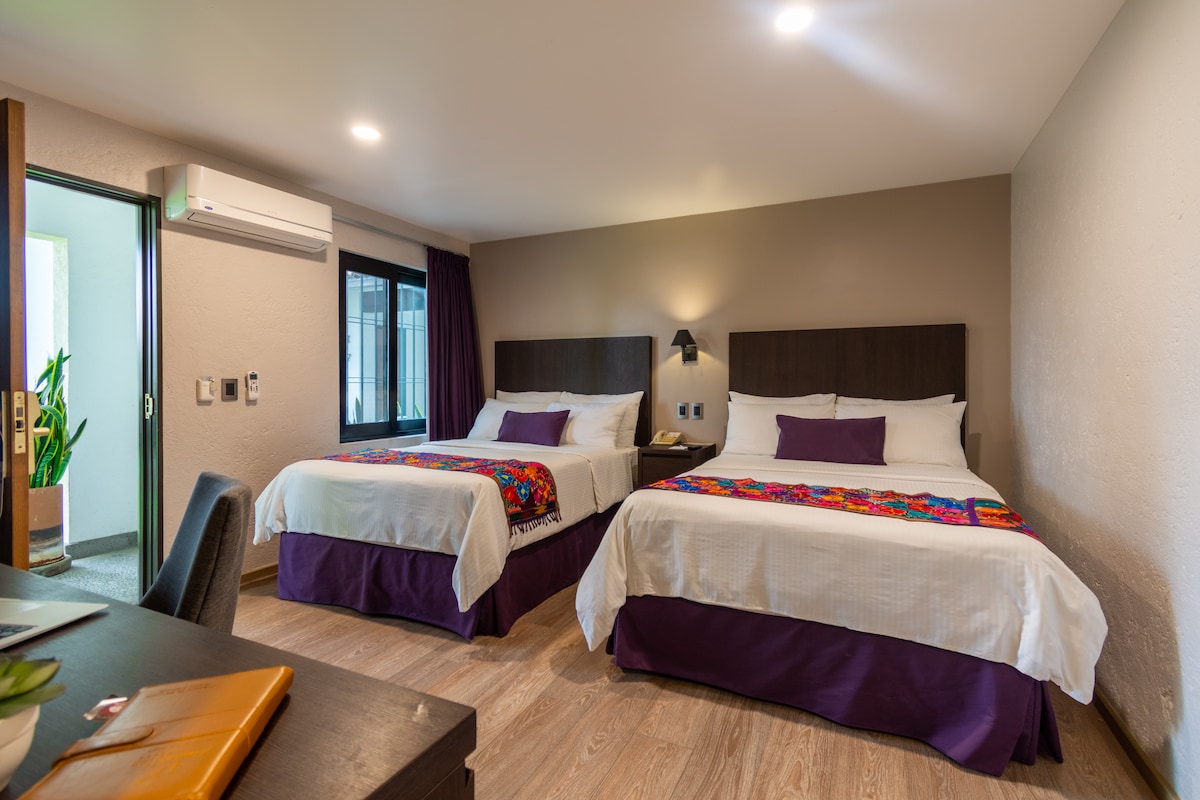 The room features two spacious beds with decorative throws at the foot. A neutral wall color complements the modern furniture, including a desk. Ample natural light enters through the window, while an air conditioning unit is mounted for comfort.