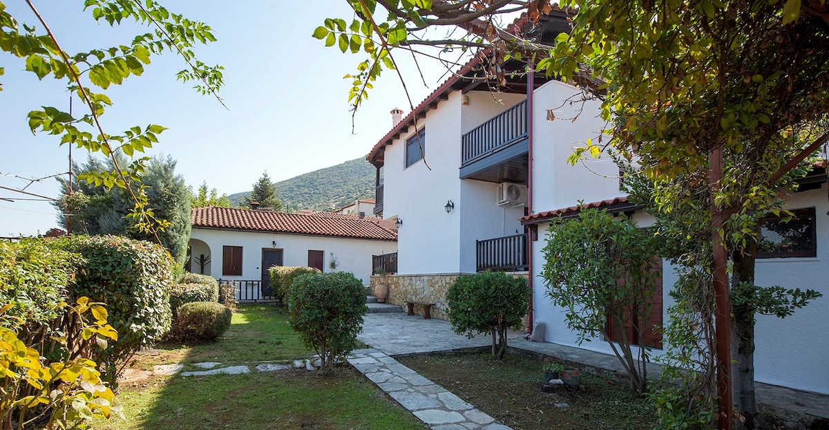 The image features two separate buildings in a garden setting. The main structure is two stories with a balcony, while a smaller building is adjacent. Well-maintained pathways and hedges create a welcoming atmosphere in the shared outdoor space.