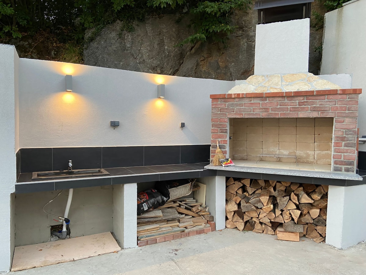 An outdoor barbecue area features a brick grill with a large stone chimney. A countertop with a sink is positioned alongside, and firewood is neatly stacked beneath. Soft, warm lighting highlights the space, set against a backdrop of natural stone and greenery.